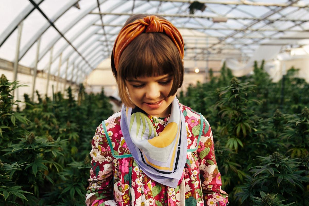 Charlotte Figi vestindo um casaco floral e lenço colorido, em uma estufa com plantas de cannabis. 