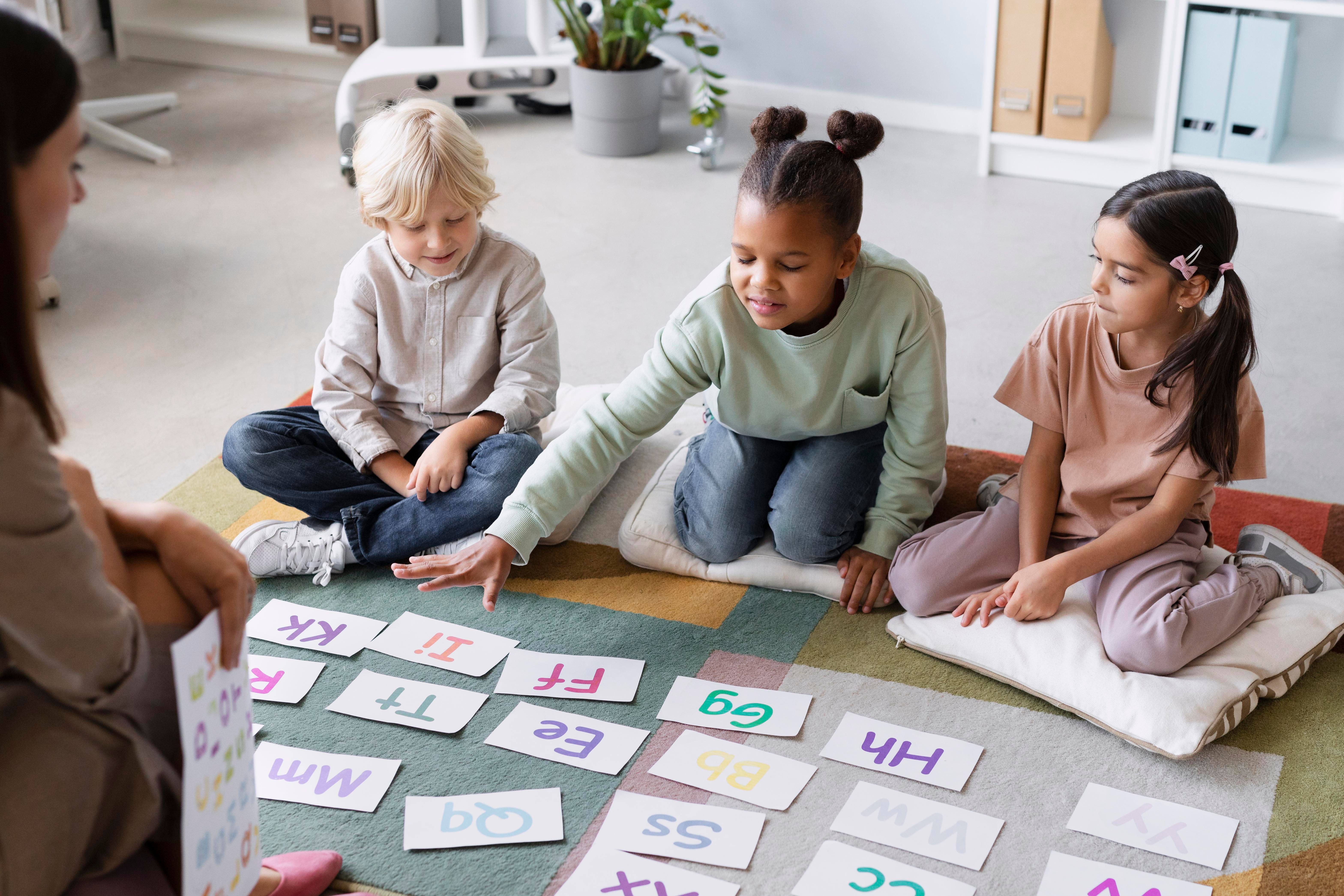 Três crianças sentadas no chão de uma sala de aula participam de uma atividade educativa com cartões coloridos contendo letras.