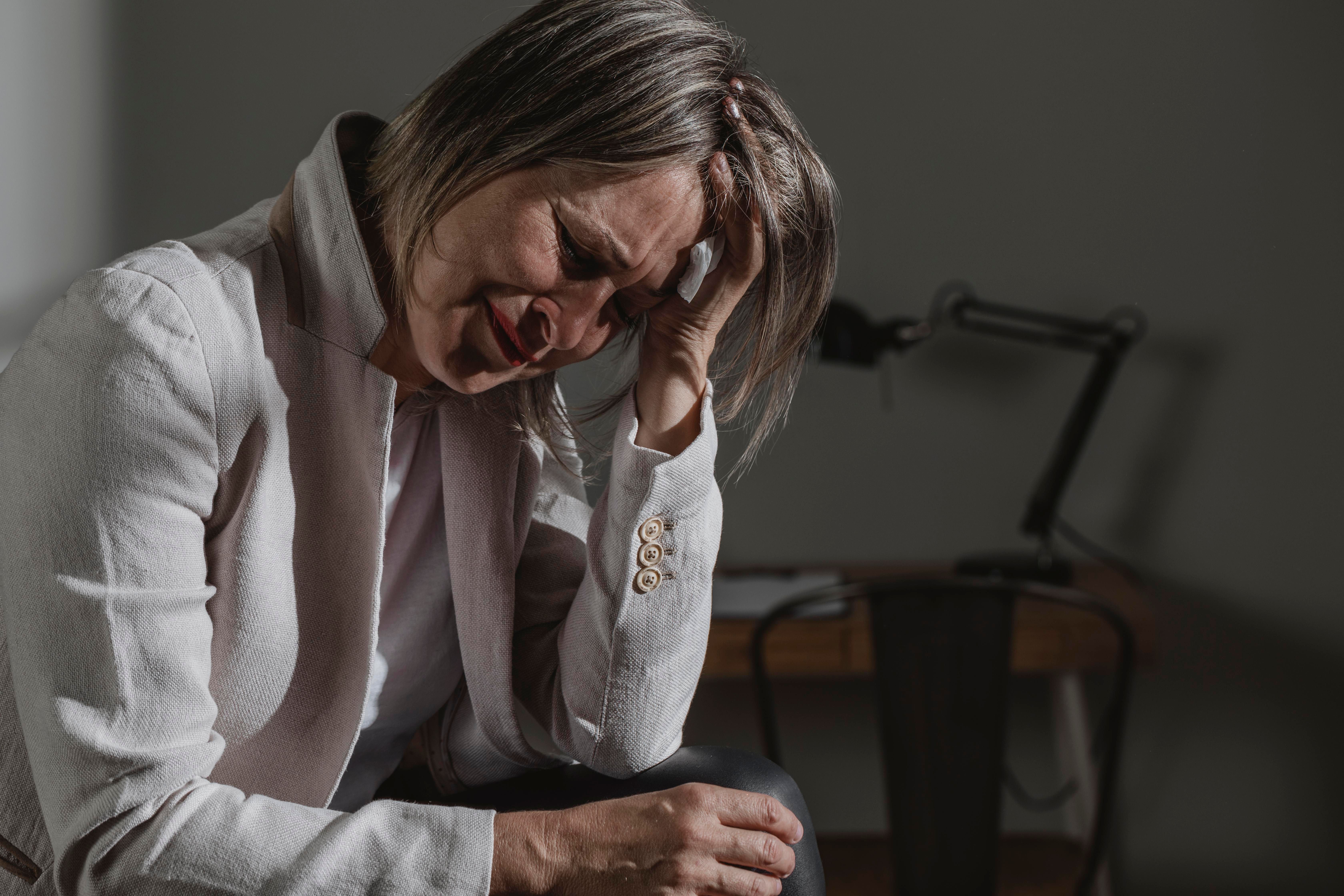 Mulher de meia-idade visivelmente abalada, sentada com a cabeça baixa e a mão na testa, indicando sofrimento psicológico. 