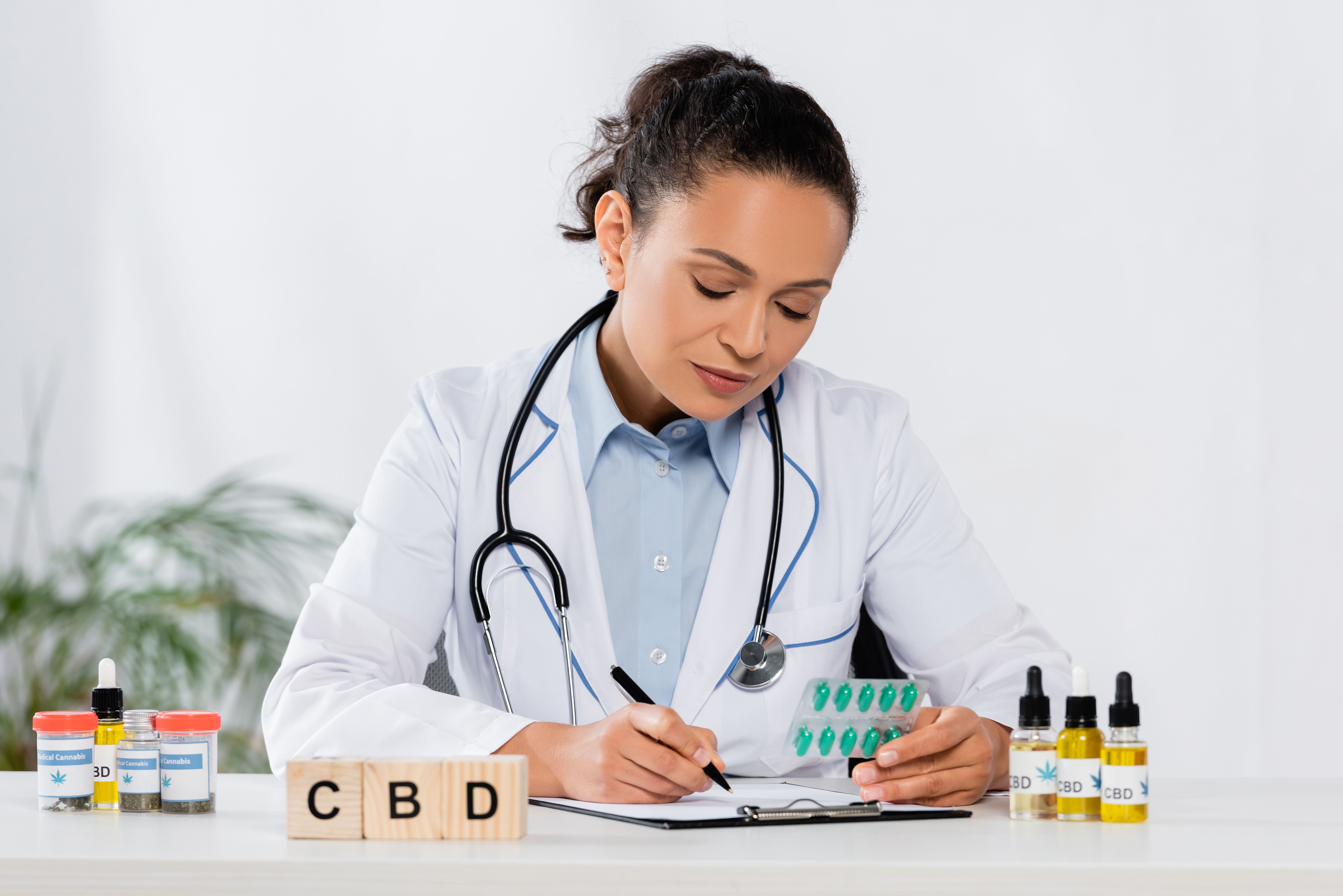 Médica analisando uma cartela de comprimidos enquanto escreve em um prontuário, sobre a mesa, há frascos de óleo de CBD 