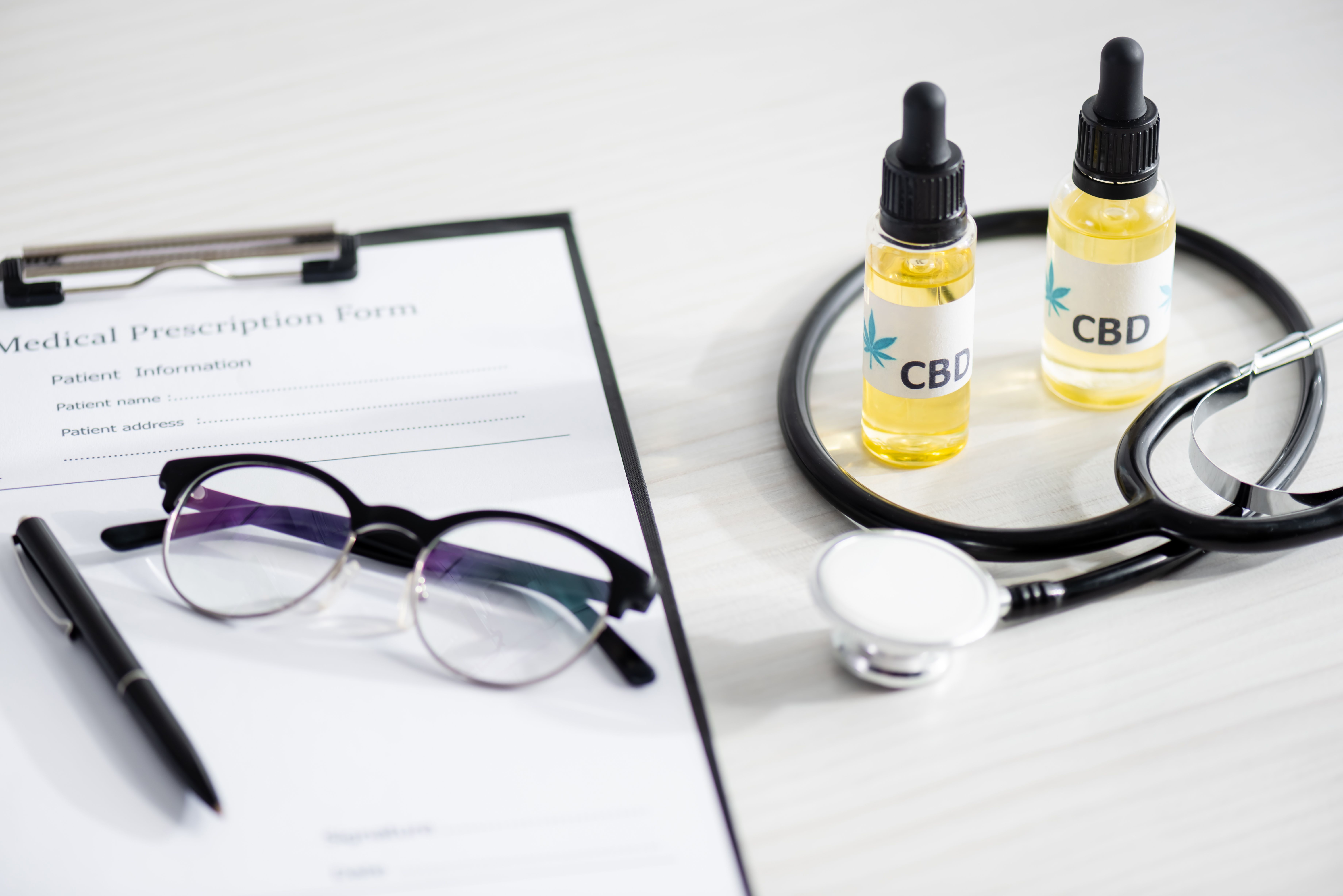 Na imagem, há duas embalagens de óleo de CBD com conta-gotas, um estetoscópio, óculos e um formulário de prescrição médica, dispostos sobre uma superfície clara.