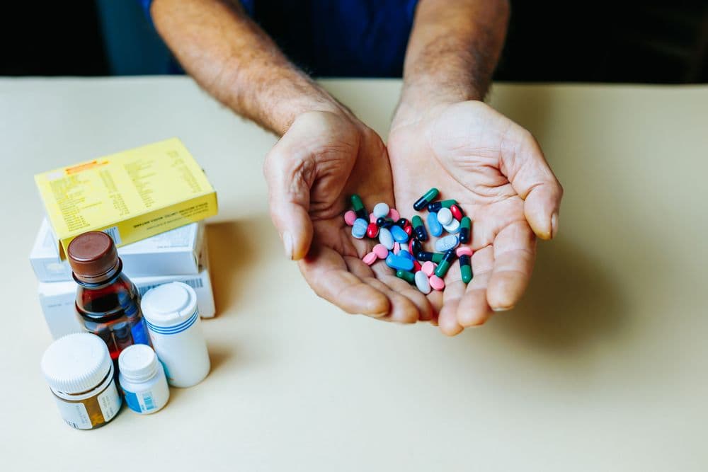 Mãos segurando cápsulas e comprimidos diversos ao lado de caixas de medicamentos.