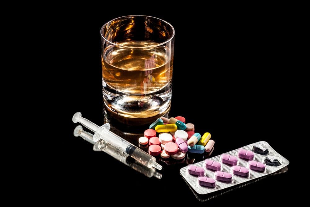 Copo de bebida alcoólica ao lado de comprimidos, seringas e cartela de remédios.