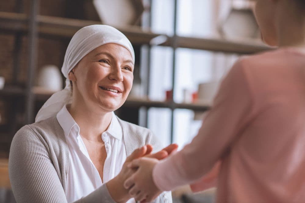 Mulher com câncer sorrindo e segurando as mãos de uma criança, transmitindo acolhimento e esperança.